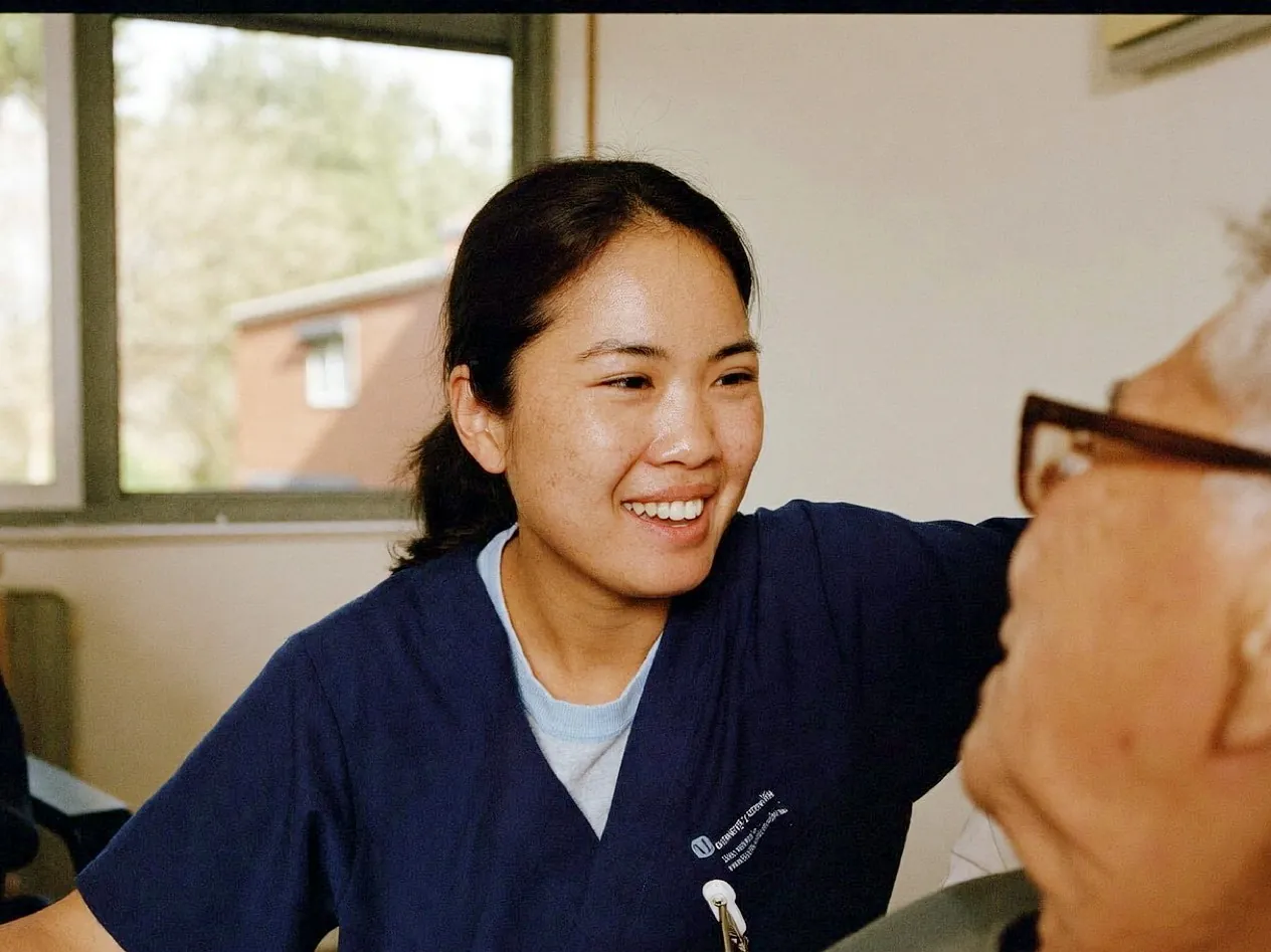 Filipina nurse in navy scrubs with an older patient on a bright hospital ward